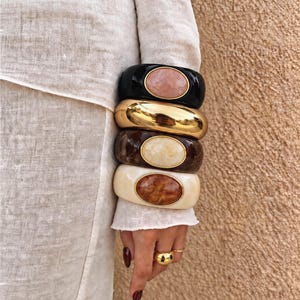 May include: A stack of four wide bangle bracelets. The top is black with a pink oval stone, the second is polished gold-tone metal, the third is brown marbled resin with a cream oval stone, and the bottom is cream resin with a brown marbled oval stone.