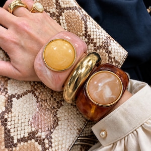May include: A stack of three wide bracelets including a pale pink resin cuff with a large yellow centre stone, a polished gold-tone metal bangle, and a dark brown tortoiseshell resin cuff with a cream-coloured centre stone.