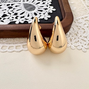 May include: A pair of polished gold-tone earrings shaped like smooth, rounded teardrops. The earrings feature a reflective, metallic surface with a curved, bulbous design that tapers to a point at the top, showcasing a solid, seamless construction.