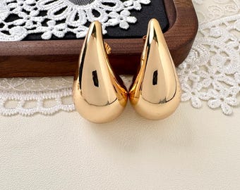 May include: A pair of polished gold-tone teardrop-shaped earrings with a smooth, reflective surface. The earrings feature a rounded, bulbous base that tapers toward the top, showcasing a solid metallic finish with visible light reflections.