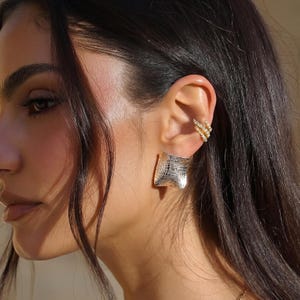 May include: A close-up of a person's ear adorned with two pieces of jewelry. A gold ear cuff with small, sparkling stones and a large, square, textured silver earring. The earring has a unique, abstract design.