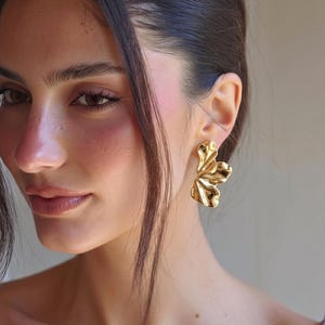 Könnte beinhalten: Goldfarbene Statement-Ohrringe mit einem strukturierten, organischen Design. Die Ohrringe haben eine fließende, blütenblattartige Form, die das Licht einfängt. Ein auffälliges Accessoire, perfekt, um jedem Outfit einen Hauch von Eleganz zu verleihen.