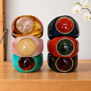 May include: Two stacks of three chunky resin rings, each featuring a smooth, circular cabochon center framed in gold-tone metal. The left stack includes amber, pale pink, and teal rings. The right stack features navy, red, and black rings.