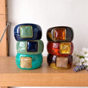 May include: Two stacks of six wide resin bangles, each featuring a central square stone set in a gold-toned metal frame. The bangles display various marbled patterns in shades of navy, sage green, turquoise, dark brown, red, and amber.