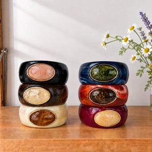 May include: Two stacks of three wide resin bangles, each featuring a central oval cabochon framed in gold-tone metal. The left stack includes black, dark brown, and cream bangles. The right stack features blue, orange, and deep purple marbled bangles.