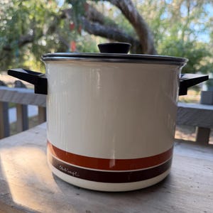May include: Cream-colored enamel pot with a black lid and handles. The pot features two horizontal stripes in shades of brown and a signature. A vintage kitchenware item.