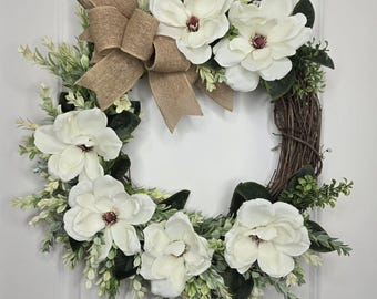 May include: A square wreath featuring large white magnolia flowers with burgundy centres, green leaves, and light green sprigs. A burlap bow is at the top. The wreath is made on a brown twig base.