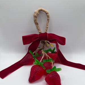May include: A decorative hanging ornament featuring a cluster of red strawberries with green stems, tied with twine and a deep red velvet bow. The ornament has a jute loop for hanging.