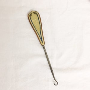 Peut inclure: Un boutonniere ancien avec une poignée décorative. La poignée est de couleur crème clair avec une bordure marron et des accents bleus. Le crochet est en métal et a une extrémité incurvée.
