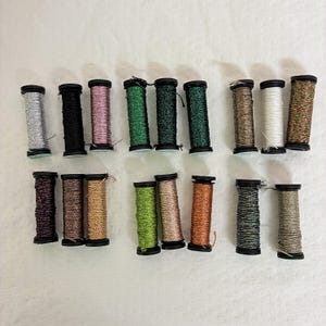 May include: A collection of spools of metallic thread in various colors, including silver, black, pink, green, brown, white, and copper. The spools are arranged in rows on a white surface, showcasing the variety of colors available for crafting and sewing projects.