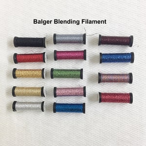 May include: A collection of spools of Balger Blending Filament in various colors, including black, silver, red, pink, gold, green, and blue. Each spool has a black rim and is filled with glittery thread. The text "Balger Blending Filament" is displayed at the top.