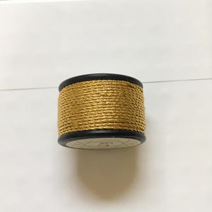 Puede incluir: Una bobina de hilo metálico dorado. El hilo está enrollado alrededor de un núcleo de plástico negro. La parte inferior de la bobina tiene una etiqueta blanca con texto y símbolos negros. El hilo tiene un aspecto brillante y texturizado.