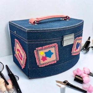 May include: A denim makeup case with a curved top and a pink handle. The case features decorative patches with star and heart designs, outlined with pink fringe. A silver clasp secures the case.