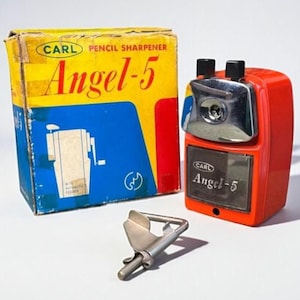 May include: A vintage red and silver pencil sharpener with the text "CARL Angel-5" and its original box. The box is yellow, blue, and red with the same text and a diagram of the sharpener. A silver blade is in front.