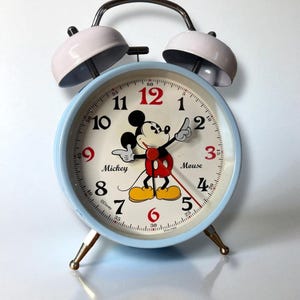 May include: A light blue Mickey Mouse alarm clock with a white face and red numbers. The clock features a cartoon image of Mickey Mouse and has two light pink bells on top. The clock is approximately 6 inches tall.