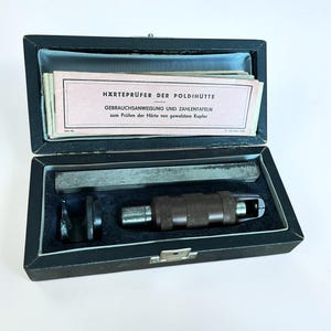 May include: A vintage Poldihütte hardness tester set housed in a black rectangular case with a blue velvet interior. The kit includes a metal bar, a dark brown knurled tool, a black component, and an instruction booklet titled Härteprüfer der Poldihütte.