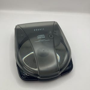 Puede incluir: Un limpiador automático de discos compactos Nuance negro. El dispositivo tiene una tapa transparente tintada y una base negra. Las palabras "Nuance" y "Compact Disc Digital Audio" están impresas en la tapa.