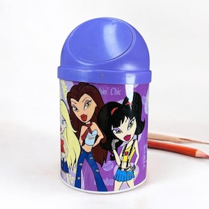 May include: A purple pencil holder with a flip-top lid. The cylindrical container features cartoon illustrations of three girls in fashionable outfits. Two pencils are visible on the right side of the holder.