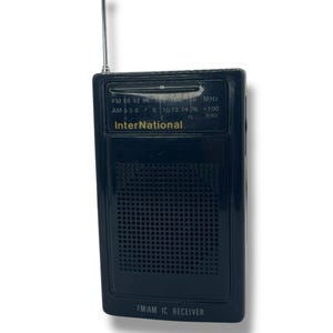 Könnte beinhalten: Ein schwarzes FM/AM IC-Empfängerradio mit Teleskopantenne. Das Radio hat ein Lautsprechergitter und ein Einstellrad mit dem Wort "International" auf der Vorderseite. Das Radio ist etwa 13 cm hoch.