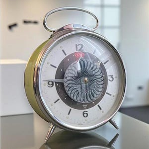 May include: A vintage alarm clock with a silver handle and legs. The clock face is white with black numerals and a decorative center. The clock's body is a light green color with a silver trim.
