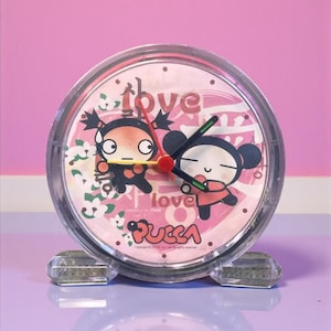 May include: A clear, round alarm clock featuring the characters Pucca and Garu, with the word "love" and the Pucca logo on the face. The clock has a pink background and a clear plastic casing. The hands are red and green.