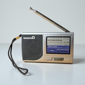 May include: A silver and gold Radisson portable radio with a retractable antenna. The radio has a black speaker grill, a digital display, and a wrist strap. The display shows the text "SLIM STYLE" and "AM & FM 2 BANE".