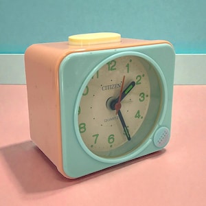May include: A vintage Citizen quartz alarm clock in pastel colours. The square clock has a peach-coloured body and a mint green face with a cream-coloured clock face. The clock face has black numbers and hands, with the word "Citizen" printed on the face.