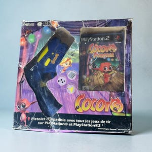 May include: A blue plastic light gun controller for PlayStation 2 in its original cardboard packaging. The box features the Cocoto Funfair game title, a red character illustration, and text stating it is compatible with all shooting games.