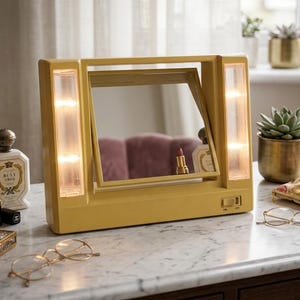 Puede incluir: Un espejo de tocador amarillo con luces integradas. El espejo es rectangular con una sección central giratoria. Un lápiz labial, frascos de perfume y gafas están sobre la superficie de mármol.