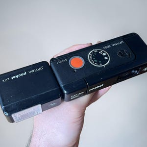 May include: A black Agfa Optima 6000 pocket camera with an attached Optima pocket LUX flash unit. The camera features a prominent orange shutter button, a circular focus dial with distance markings, and the text Optima 6000 and pocket printed.
