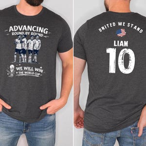May include: Dark gray t-shirt with a soccer theme. The front features the text "Advancing Round by Round" and "We Will Win The World Cup" with a team graphic. The back displays "United We Stand", the name "Liam", and the number "10".