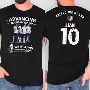 May include: Black t-shirt with a soccer team graphic. The front features a team image and text: "ADVANCING ROUND BY ROUND" and "WE WILL WIN THE WORLD CUP." The back displays "UNITED WE STAND," the name "LIAM," and the number "10."