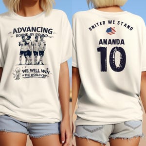 May include: White t-shirt with a soccer-themed graphic. The front features a team illustration and text: "ADVANCING ROUND BY ROUND" and "WE WILL WIN THE WORLD CUP." The back displays "UNITED WE STAND," the name "AMANDA," and the number "10."