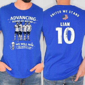 May include: Royal blue t-shirt with a soccer theme. The front features the text "ADVANCING ROUND BY ROUND" above a graphic of soccer players and a trophy. The back displays "UNITED WE STAND", the name "LIAM", and the number "10".