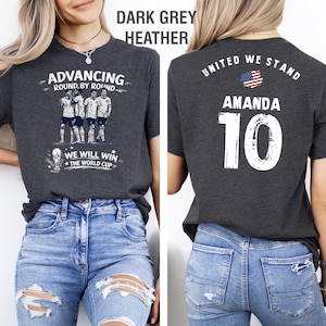 May include: Dark grey heather t-shirt with soccer-themed graphics. The front features a team illustration and text: "ADVANCING ROUND BY ROUND" and "WE WILL WIN THE WORLD CUP." The back displays "UNITED WE STAND," the name "AMANDA," and the number "10."