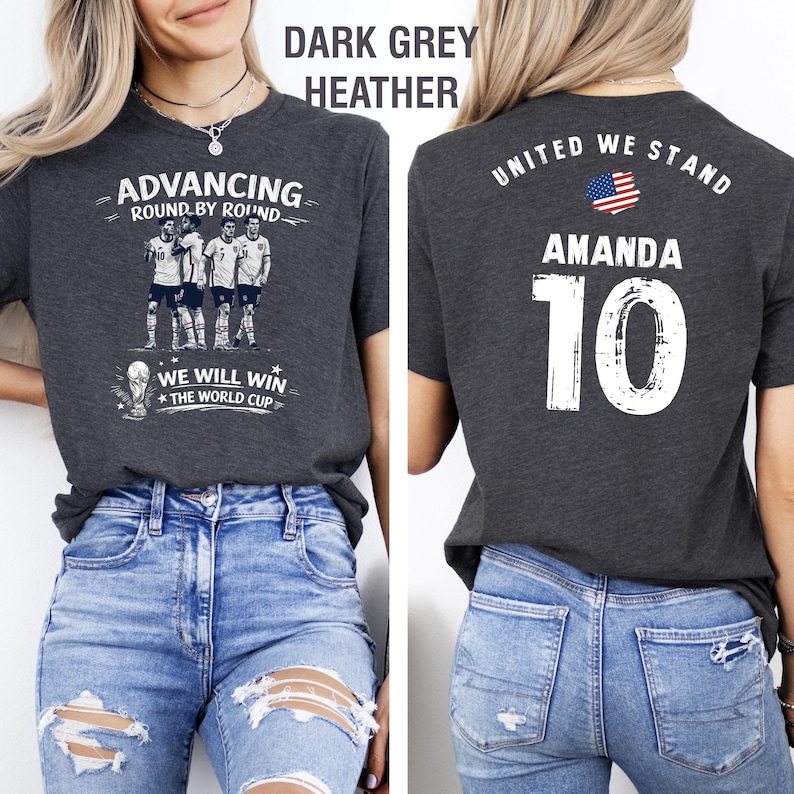 May include: Dark grey heather t-shirt with soccer-themed graphics. The front features a team illustration and text: "ADVANCING ROUND BY ROUND" and "WE WILL WIN THE WORLD CUP." The back displays "UNITED WE STAND," the name "AMANDA," and the number "10."