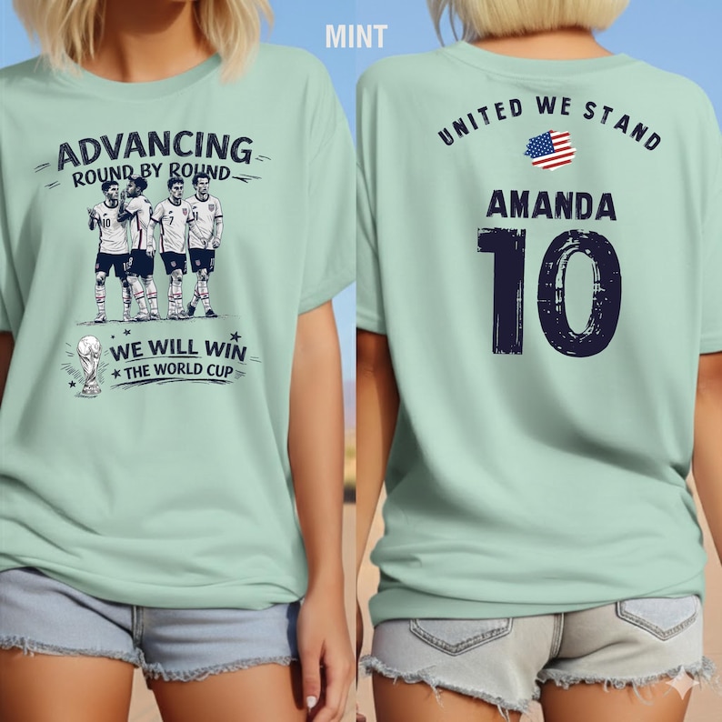 May include: Mint green t-shirt with a soccer theme. The front features a graphic of soccer players and the text "ADVANCING ROUND BY ROUND" and "WE WILL WIN THE WORLD CUP". The back displays "UNITED WE STAND", the name "AMANDA", and the number "10".