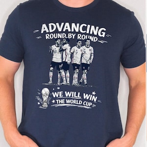 May include: Navy blue t-shirt with a graphic of four soccer players and the text "Advancing Round by Round" and "We Will Win The World Cup". The design is in white. The shirt is short-sleeved.