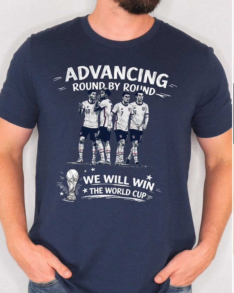 May include: Navy blue t-shirt with a graphic of four soccer players and the text "Advancing Round by Round" and "We Will Win The World Cup". The design is in white. The shirt is short-sleeved.