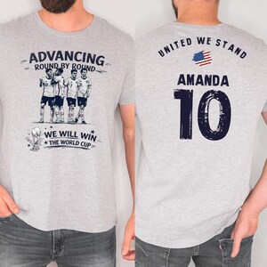 May include: Light gray t-shirt with a soccer theme. The front features a team graphic and the text "ADVANCING ROUND BY ROUND" and "WE WILL WIN THE WORLD CUP". The back displays "UNITED WE STAND", the name "AMANDA", and the number "10".