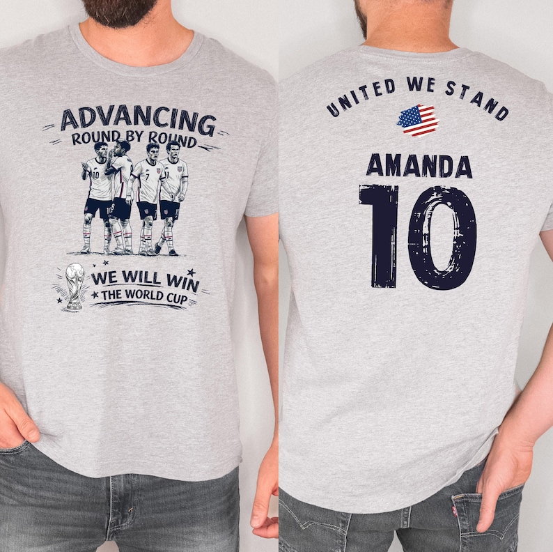 May include: Light gray t-shirt with a soccer theme. The front features a team graphic and the text "ADVANCING ROUND BY ROUND" and "WE WILL WIN THE WORLD CUP". The back displays "UNITED WE STAND", the name "AMANDA", and the number "10".