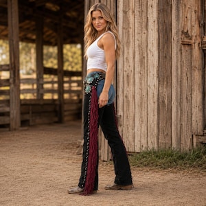 May include: Black leather chaps featuring long, deep red fringe along the outer legs and a wide, decorative belt section adorned with large, circular turquoise-colored stones and silver-toned metal studs, worn over blue denim jeans.