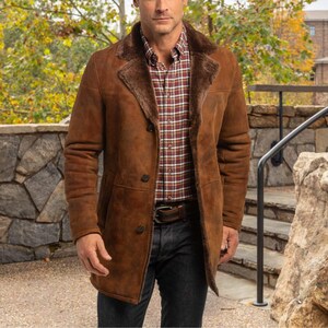 May include: A brown suede coat with a shearling collar and lining. The coat has a button closure and a classic design. The person is wearing a plaid shirt and dark jeans. The coat is a warm, stylish outerwear option.