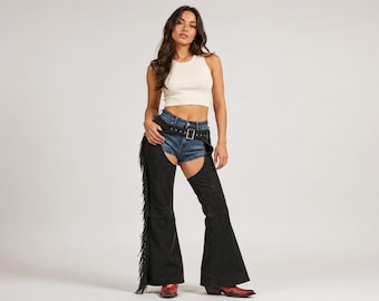May include: A white sleeveless ribbed tank top, blue denim shorts, and black leather chaps featuring long fringe along the outer legs. The chaps are secured with a black leather belt with a silver buckle, worn over the shorts, paired with red boots.
