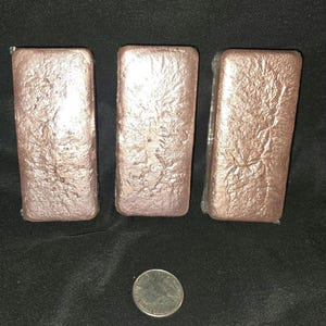 May include: Three rectangular, copper-colored metal bars with textured surfaces are arranged on a black background. A silver coin is placed near the bottom of the image for size comparison. The bars appear to be ingots.