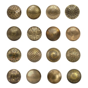 May include: A collection of sixteen round, metallic cabinet knobs arranged in a four-by-four grid. Each knob features a distinct embossed pattern in an antique brass finish, including geometric, floral, tree, and textured designs.