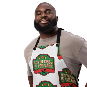 以下が含まれることがあります： 「KISS THE COOK... IF YOU DARE」のテキストが緑と赤のバナーで繰り返されるデザインの白いエプロン。エプロンには黒いストラップとボタンが付いています。デザインには、ハート、炎、交差した調理器具が含まれています。