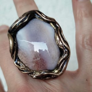 Puede incluir: Un anillo con una piedra grande, lisa y de forma ovalada con una textura superficial en tonos lavanda pálido y blanco. La piedra está engastada en una montura metálica oscura y texturizada que recuerda a ramas o vides retorcidas con reflejos bronce.