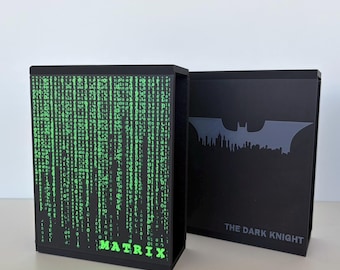 May include: Two black rectangular storage cases. The left case features a bright green digital rain pattern and the word MATRIX in green text. The right case displays a grey silhouette of a bat symbol over a city skyline with THE DARK KNIGHT text.