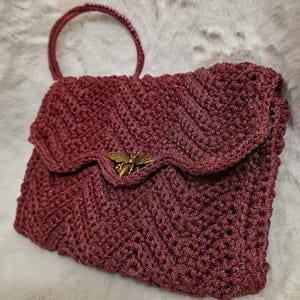 Può includere: Una borsa a mano all'uncinetto bordeaux con manico e chiusura a patta. La borsa presenta una chiusura decorativa a forma di ape color oro. Il motivo all'uncinetto è un design a chevron strutturato e la borsa ha un aspetto lucido e scintillante.